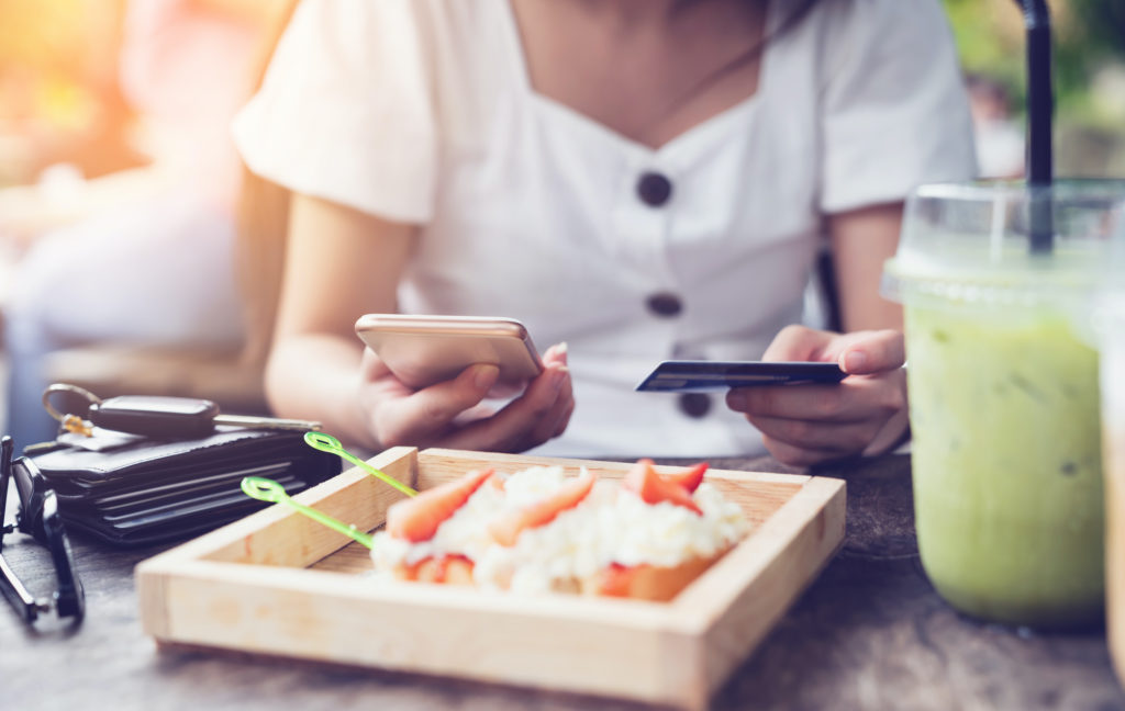 Young woman making payment through mobile smart phone with credit card while shopping online in restaurant